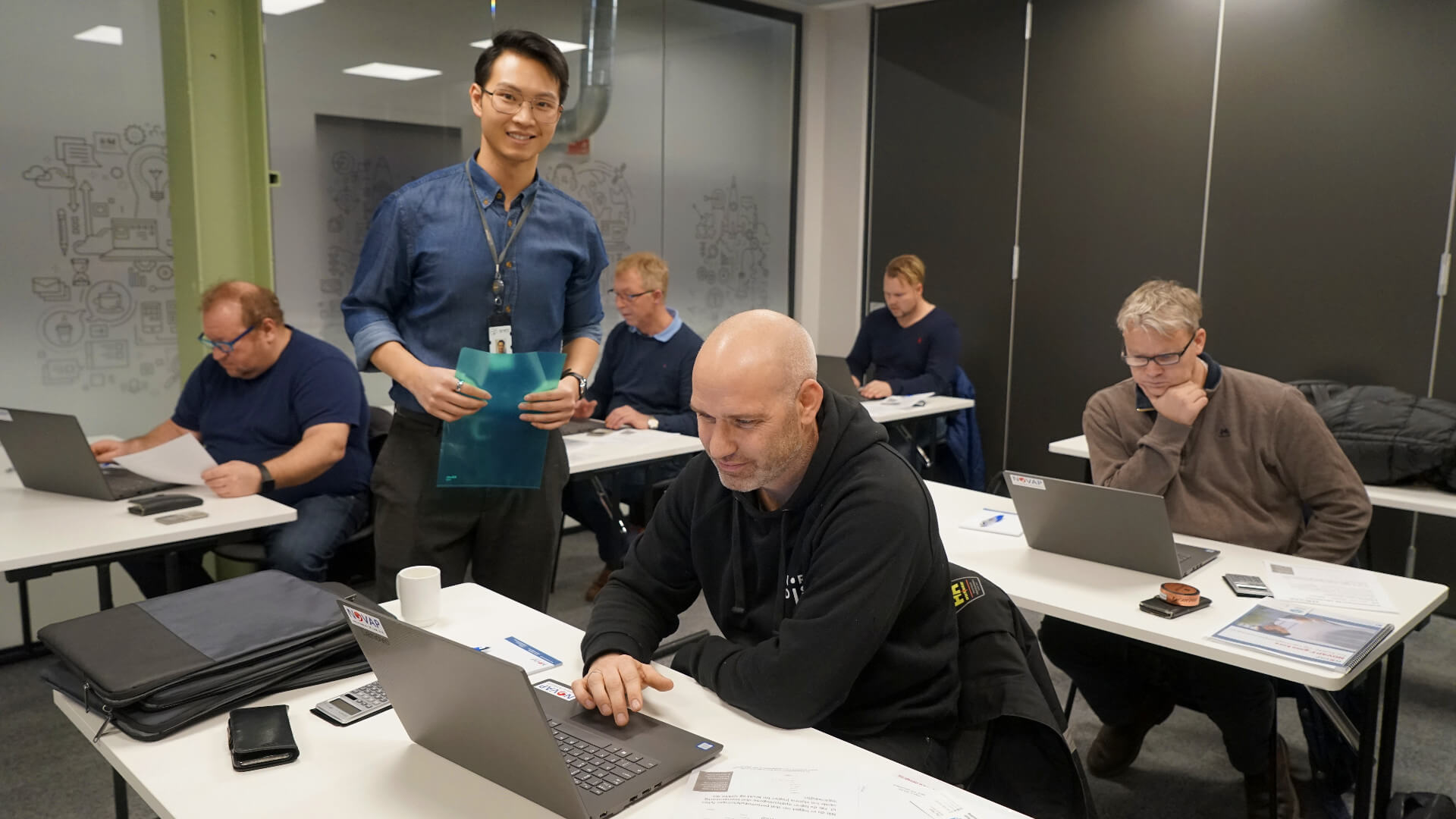 Fire personer med pc under f-gass-eksamen, med eksamensvakt John Love Phan stående mellom dem.